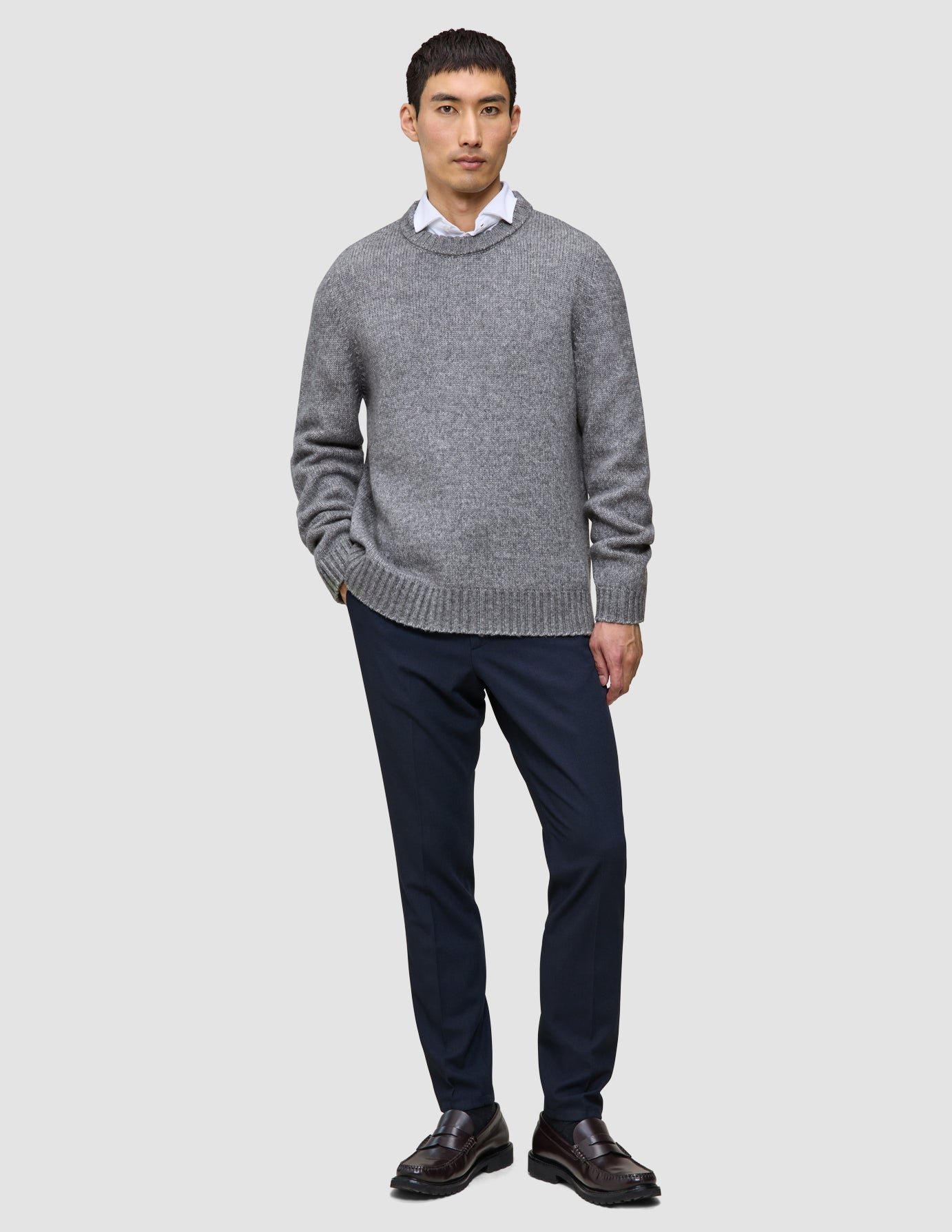 A man wearing a Chunky Soft Wool Knit Crewneck Glacier Grey sweater over a white collared shirt, dark trousers, and dark loafers.