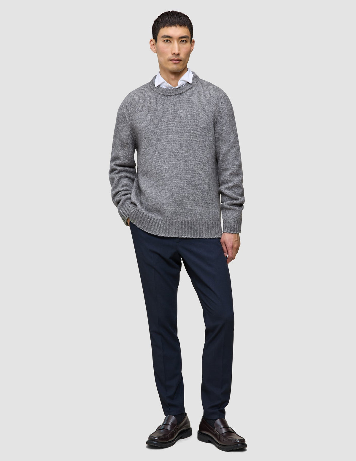 A man wearing a Chunky Soft Wool Knit Crewneck Glacier Grey sweater over a white collared shirt, dark trousers, and dark loafers.
