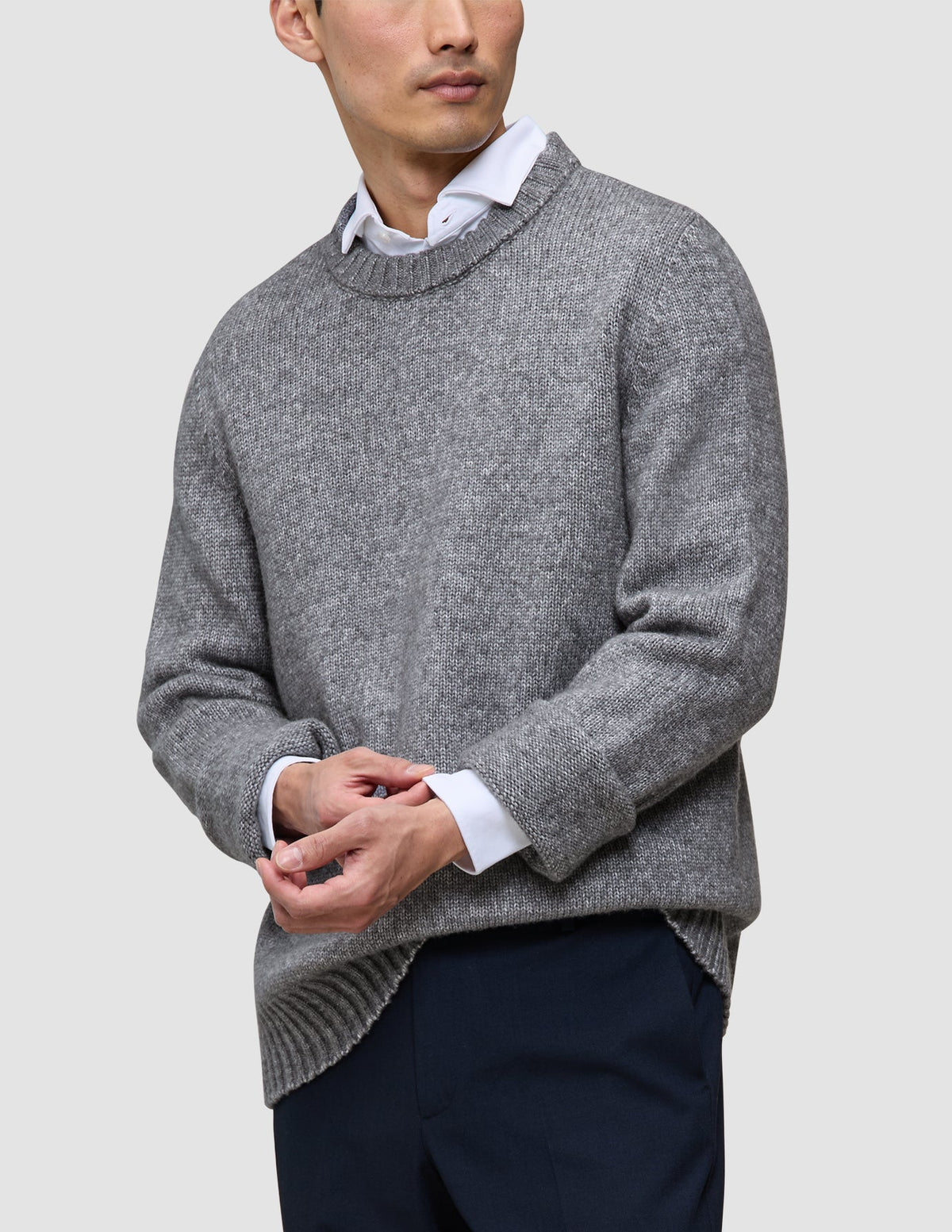 A person wearing a Chunky Soft Wool Knit Crewneck Glacier Grey sweater over a white collared shirt and dark trousers.