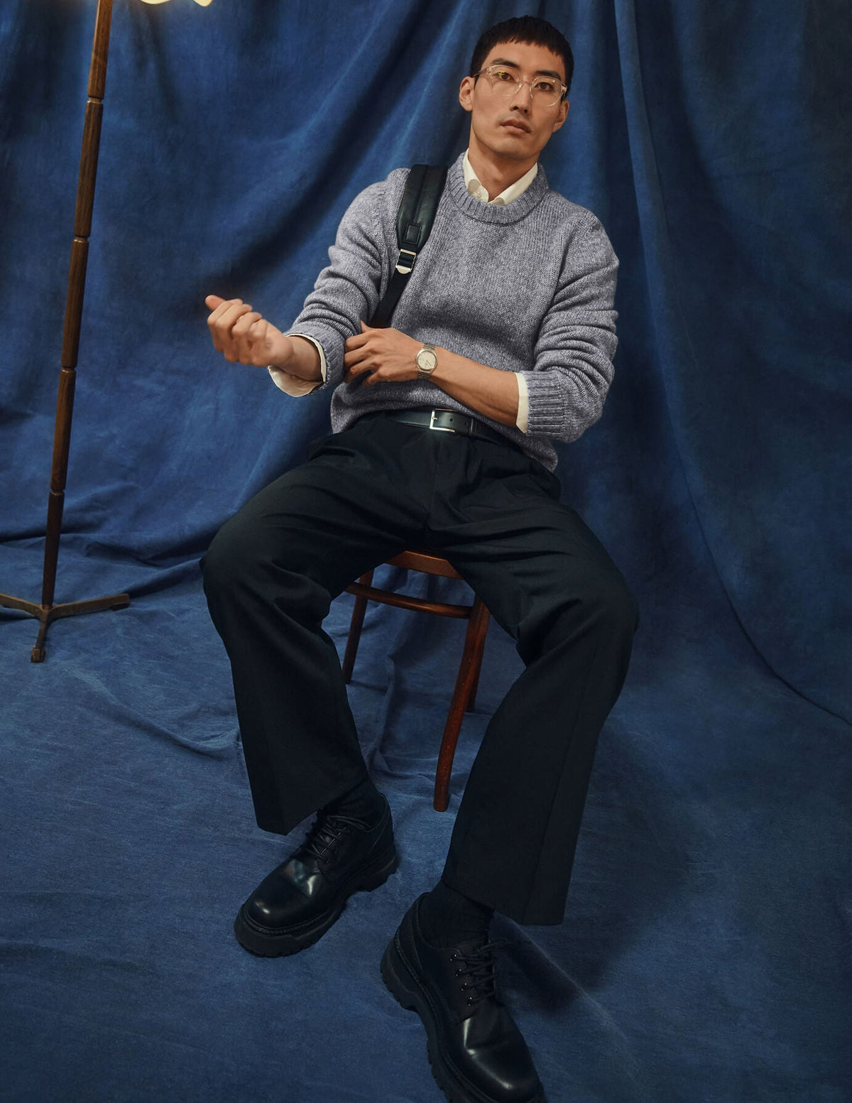A man wearing a Chunky Soft Wool Knit Crewneck Glacier Grey sweater over a collared shirt, black trousers, and black shoes, sitting on a wooden chair.