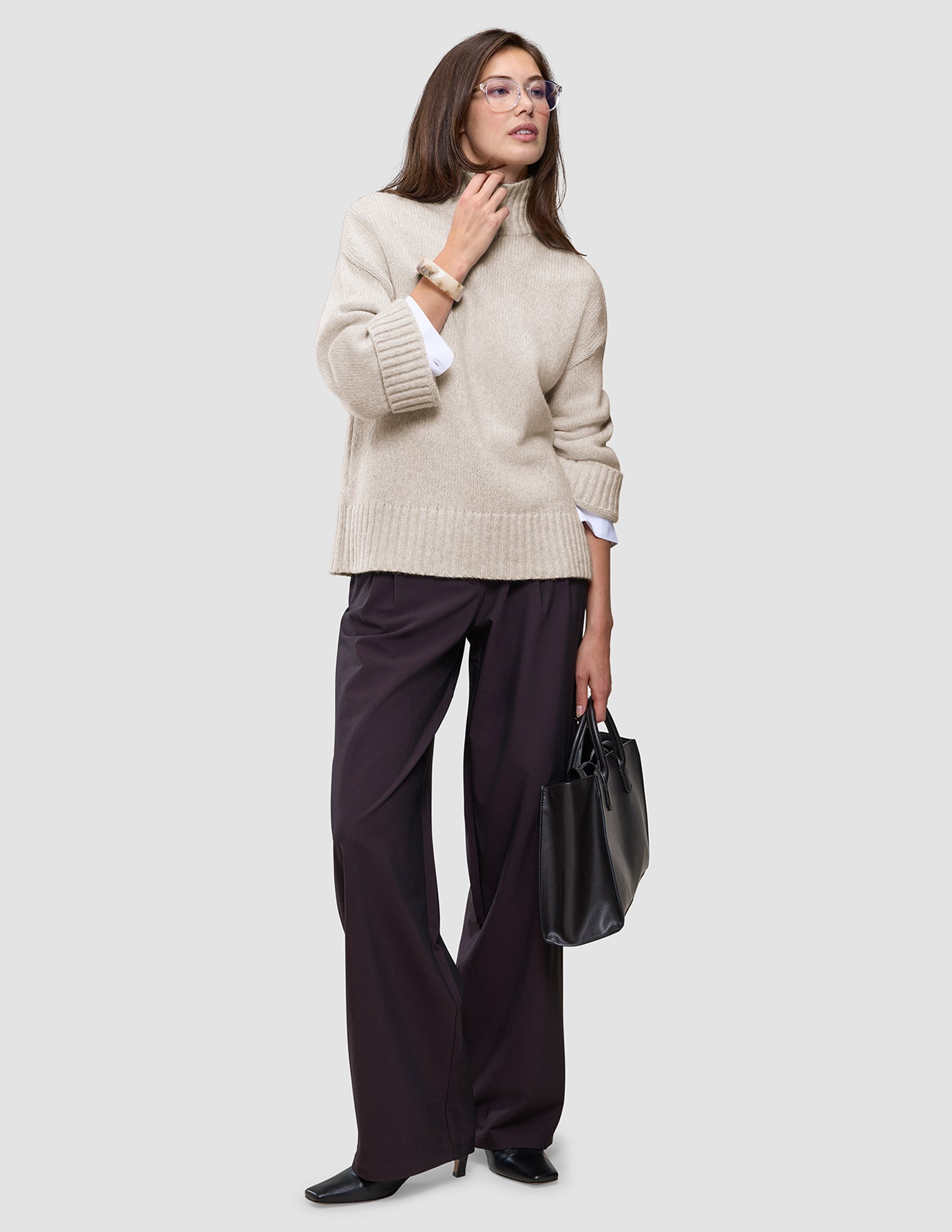A woman wearing a Women’s Chunky Soft Wool Knit Turtleneck Sugar Melange in beige with wide-leg dark trousers, carrying a black handbag.