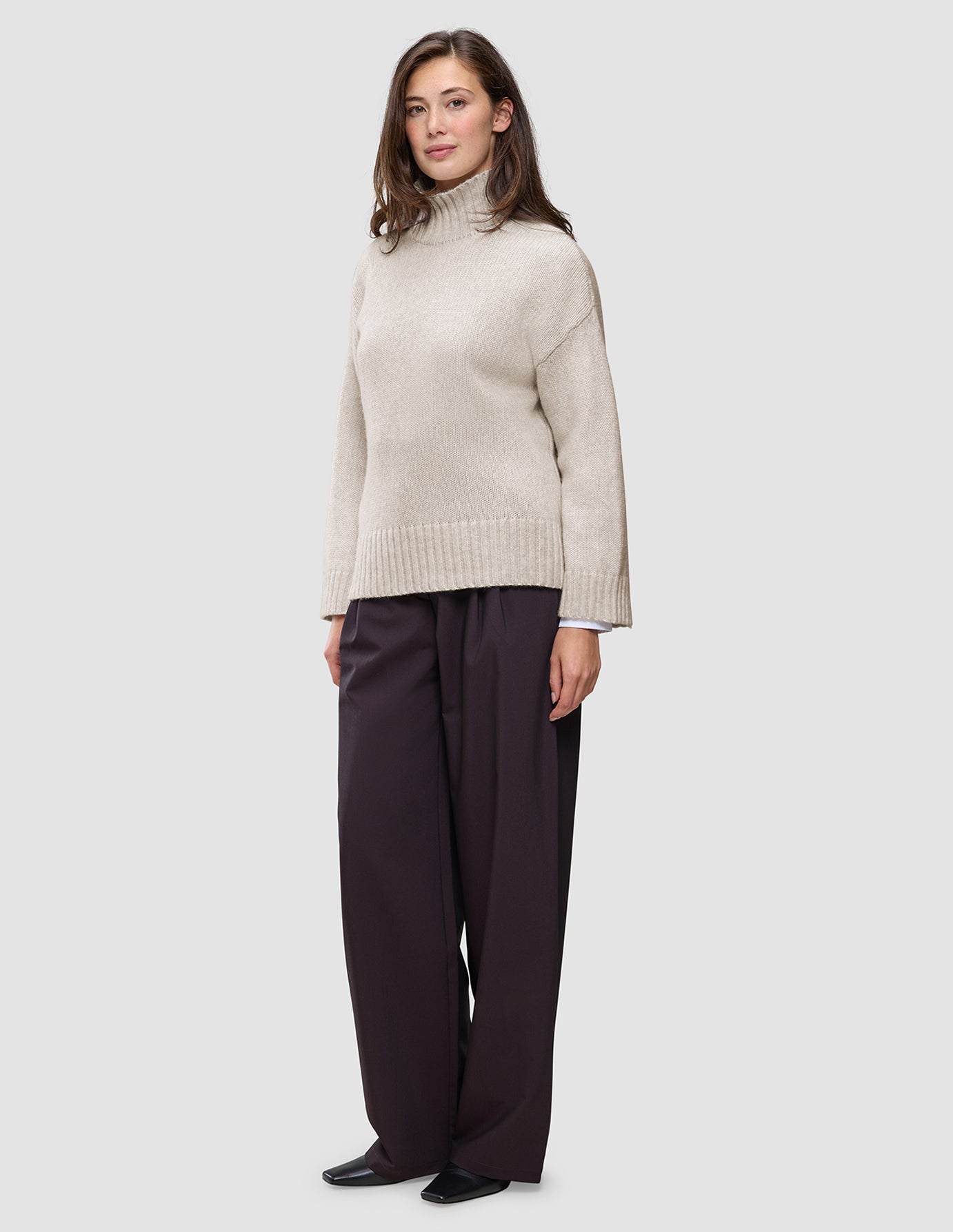 A woman wears a Women’s Chunky Soft Wool Knit Turtleneck Sugar Melange in beige with wide-leg dark trousers.