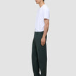 A person in profile wearing a white t-shirt, dark brown loafers, and Ease Pants Relaxed Fit Northern Pine, which are exceptionally soft, dark green, and relaxed-fit.