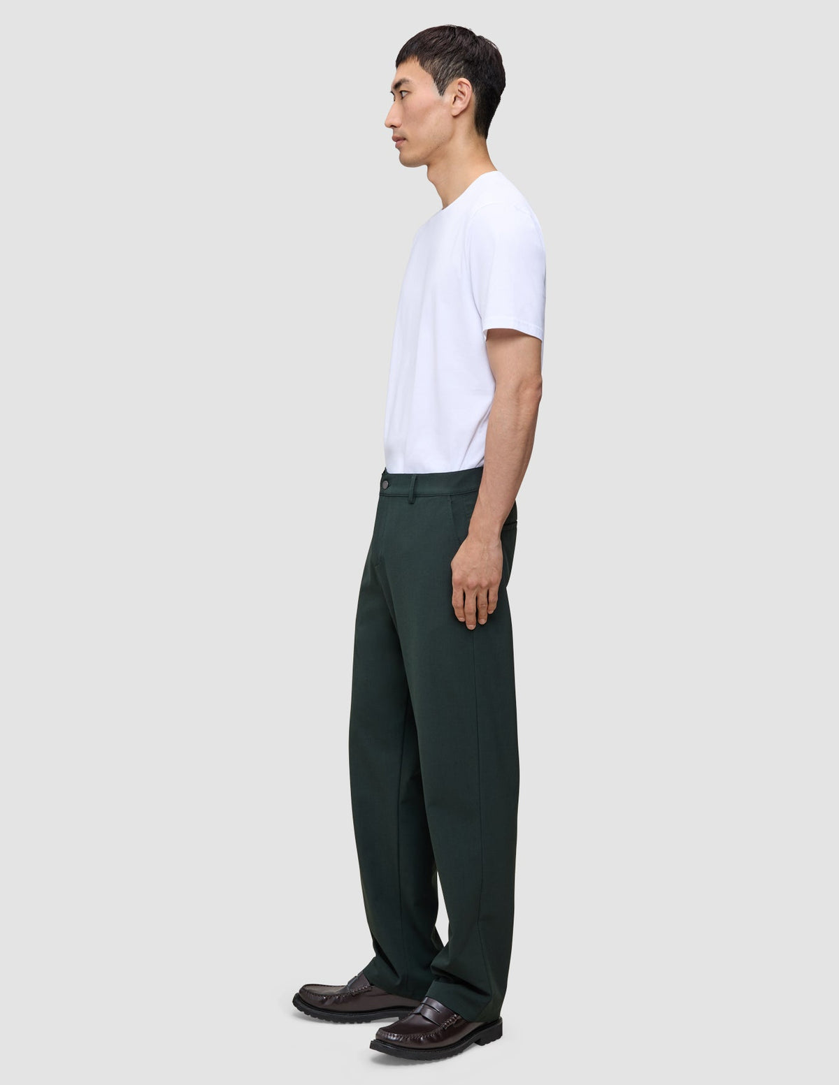 A person in profile wearing a white t-shirt, dark brown loafers, and Ease Pants Relaxed Fit Northern Pine, which are exceptionally soft, dark green, and relaxed-fit.