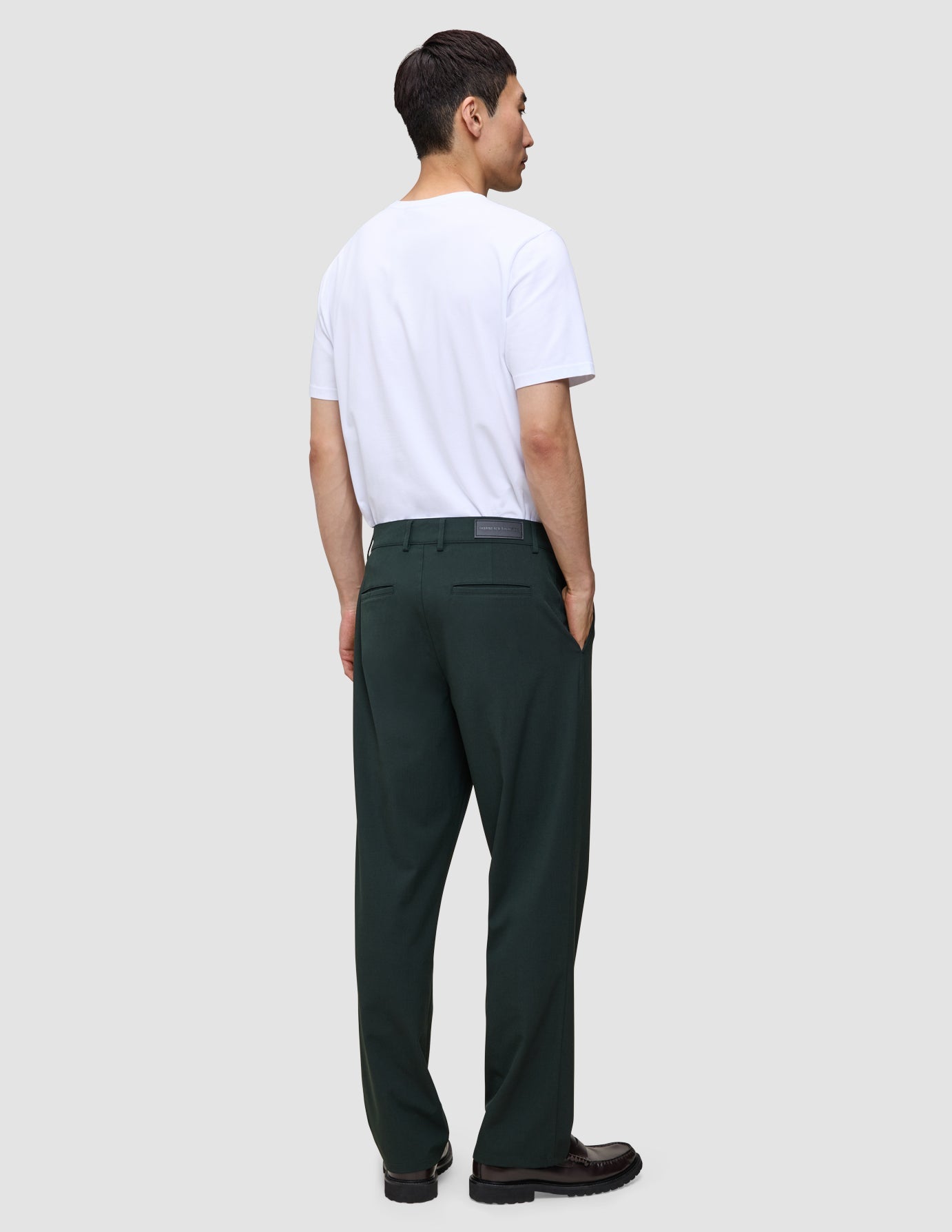 A man viewed from behind, wearing a white t-shirt and dark green Ease Pants Relaxed Fit Northern Pine with a tailored look, hands tucked into his pockets.