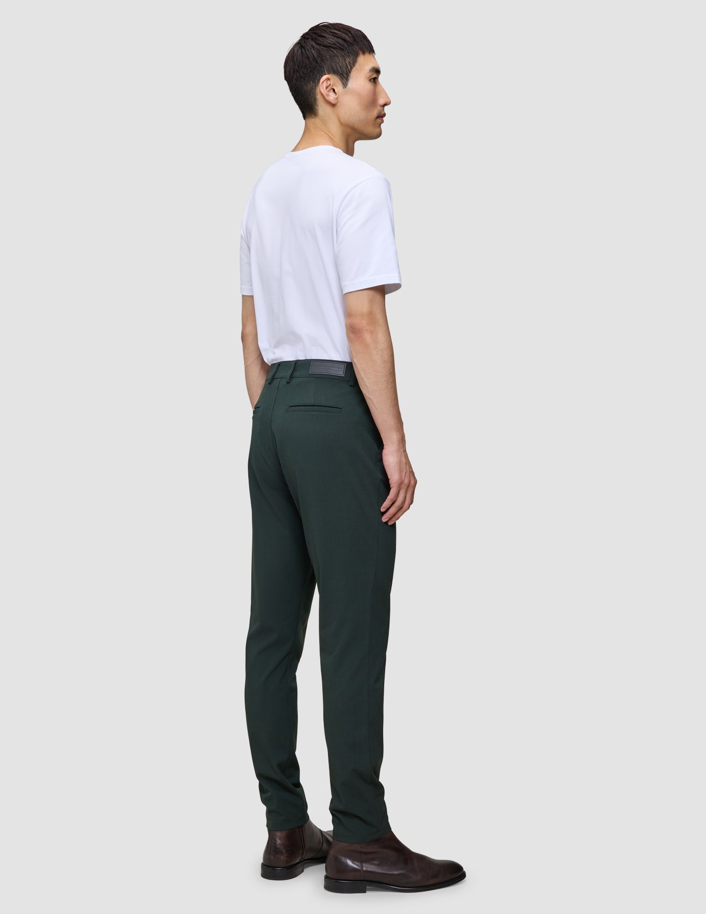 A person viewed from behind, wearing a white t-shirt, Ease Pants Slim Northern Pine, and dark leather boots.