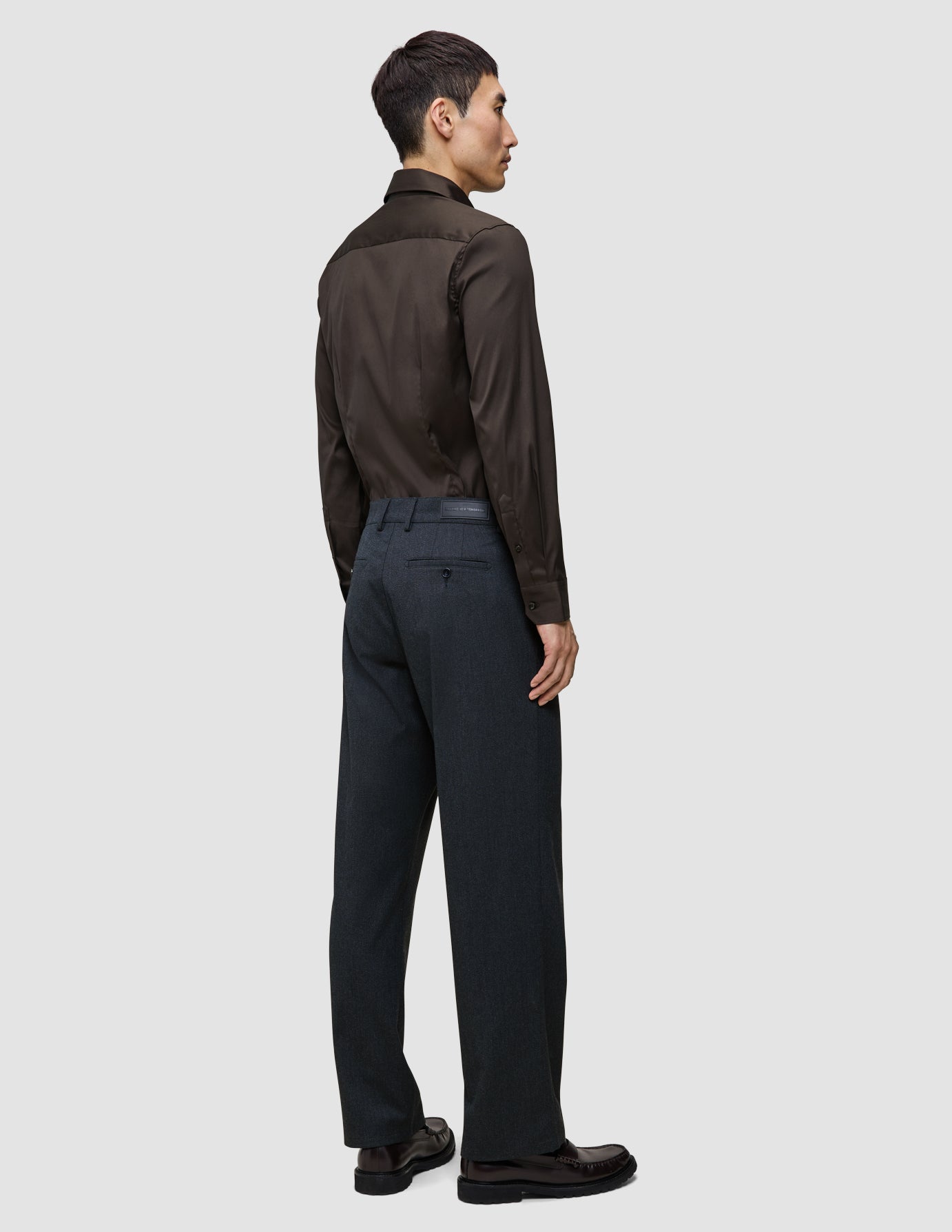 A man seen from behind wearing an Elevated Shirt Regular Dark Brown tucked into charcoal trousers and dark loafers.