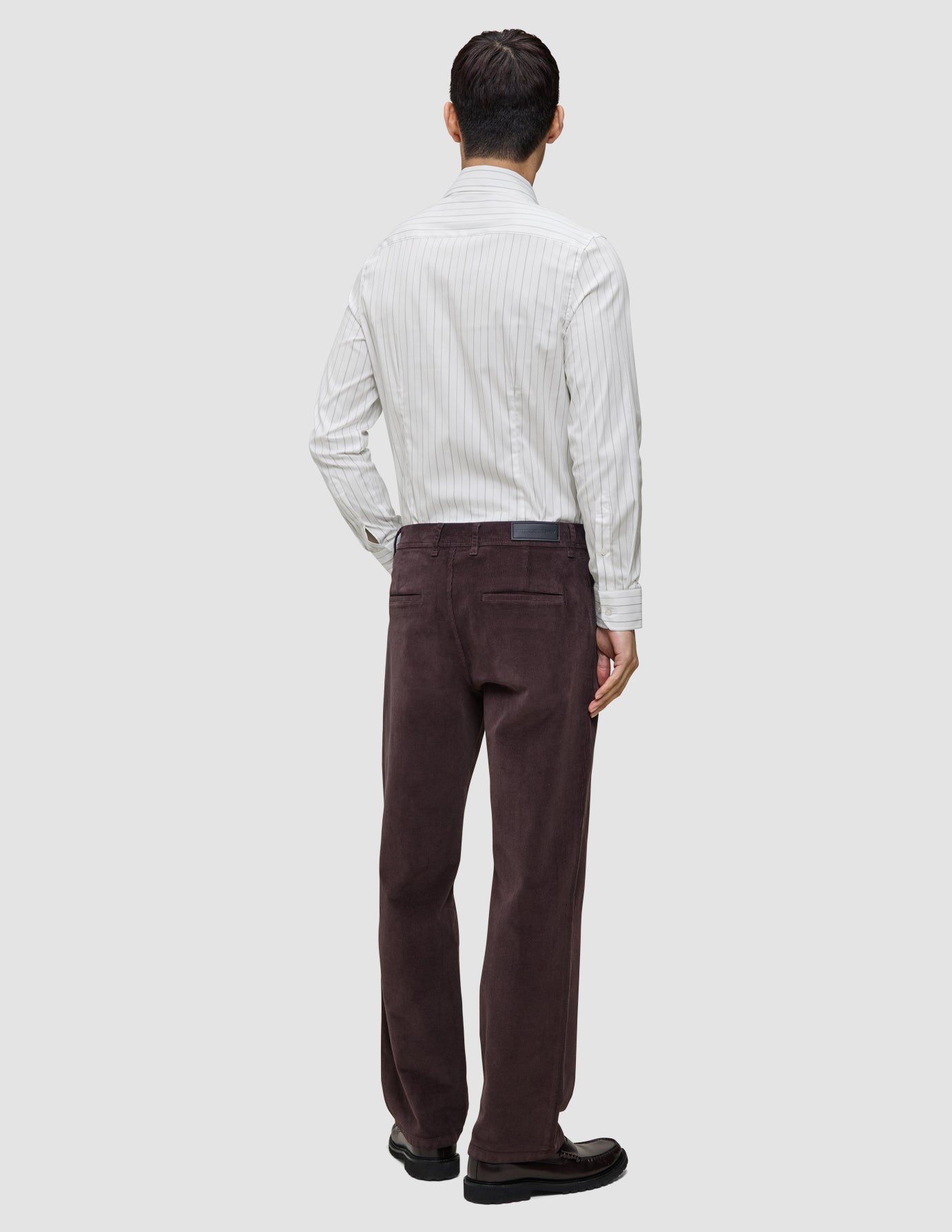 A man seen from behind wearing an Elevated Shirt Regular Light Grey Striped button-down shirt tucked into dark brown trousers and dark leather shoes.