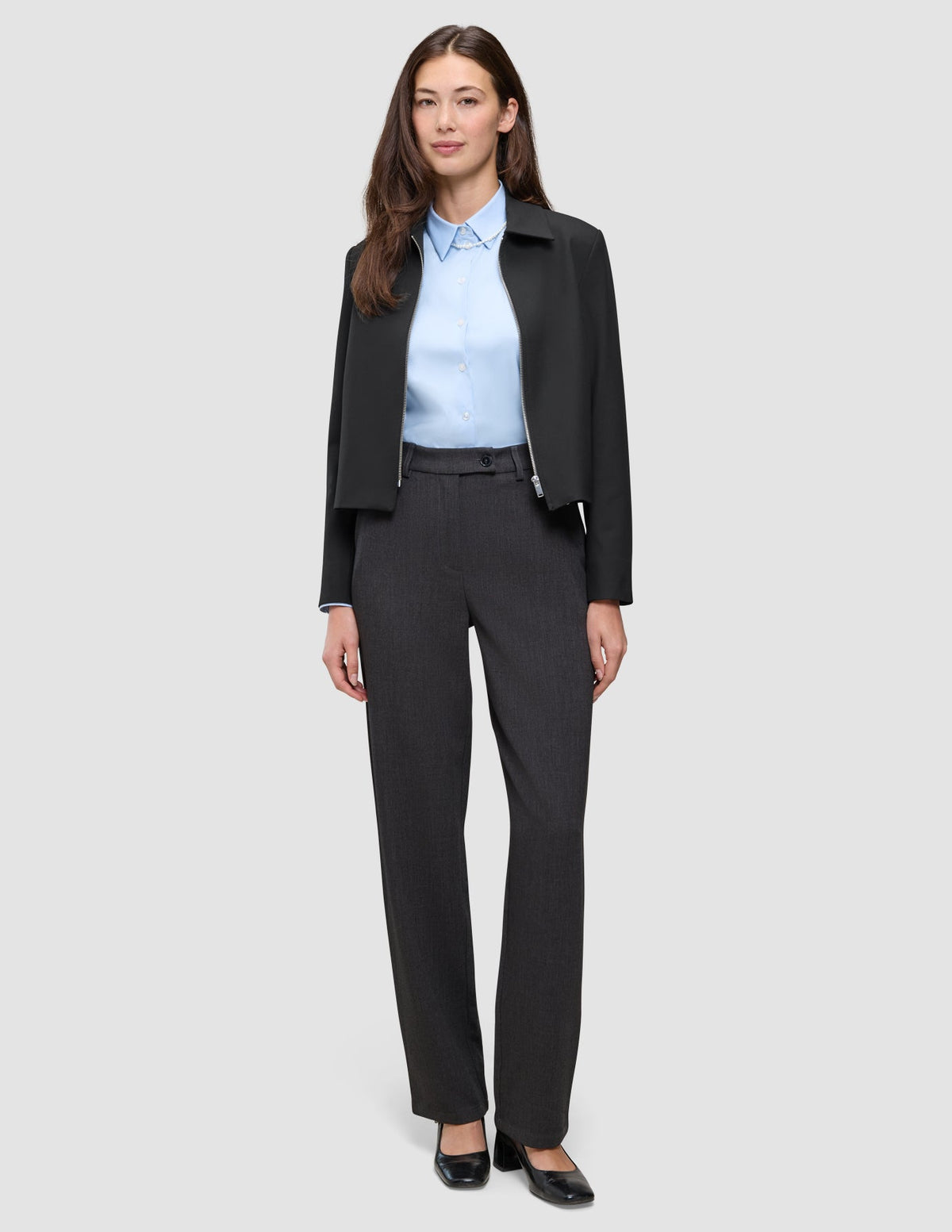 A woman wears an Essential Short Zip Jacket Black over a light blue collared shirt and dark grey trousers, paired with black block-heeled shoes.