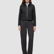 A woman wearing an Essential Short Zip Jacket Black over a light blue collared shirt, paired with charcoal grey trousers and black heels.