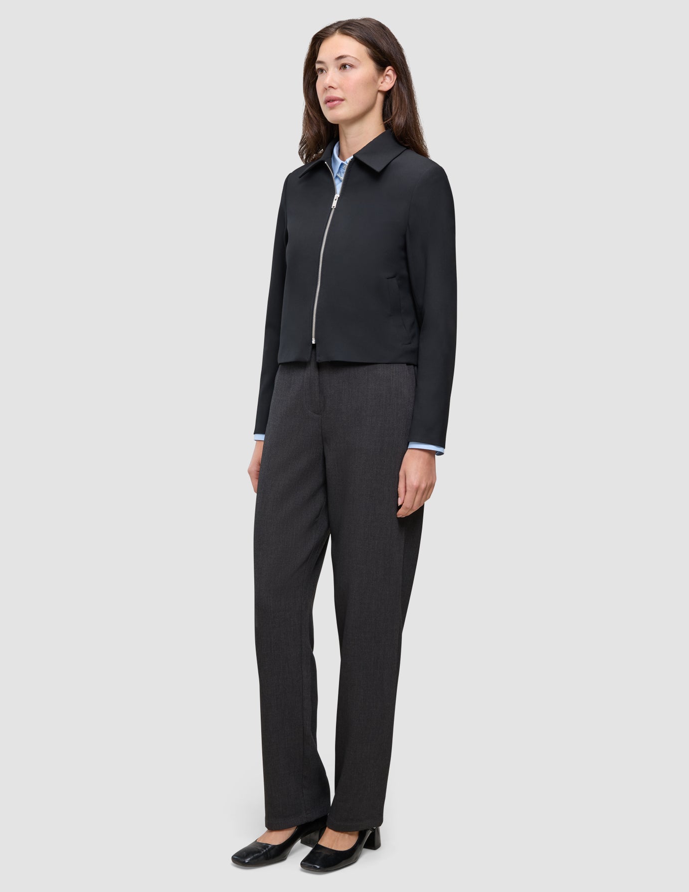 A person wears a minimalist, black Essential Short Zip Jacket over a light blue collared shirt and dark trousers.