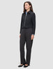 A person wears a minimalist, black Essential Short Zip Jacket over a light blue collared shirt and dark trousers.