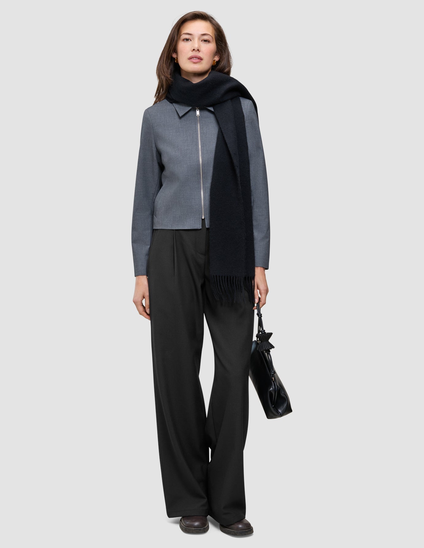 A person wearing an Essential Short Zip Jacket Dark Grey Melange, a black scarf, and black trousers, holding a black handbag.