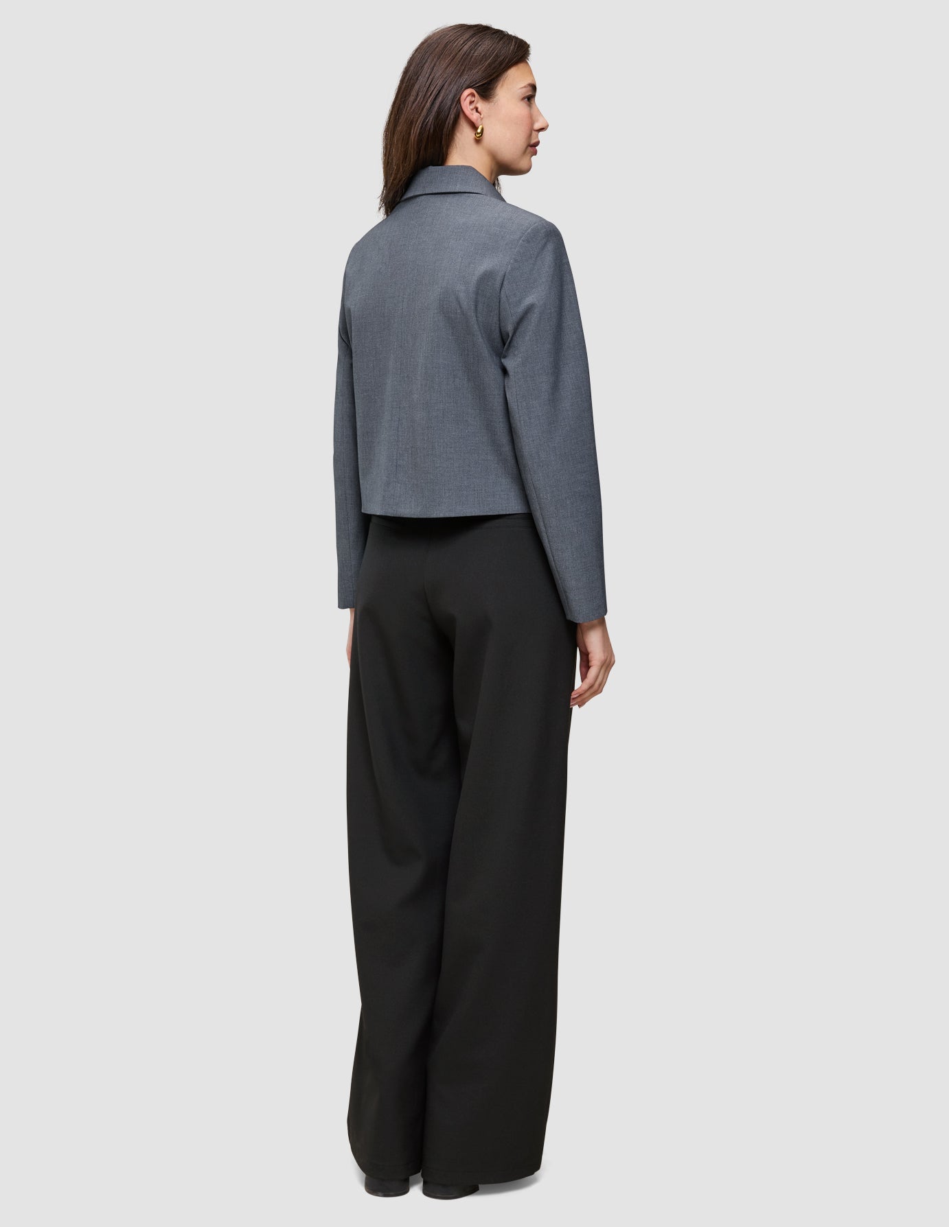 A woman viewed from behind, wearing an Essential Short Zip Jacket Dark Grey Melange and black wide-leg trousers.