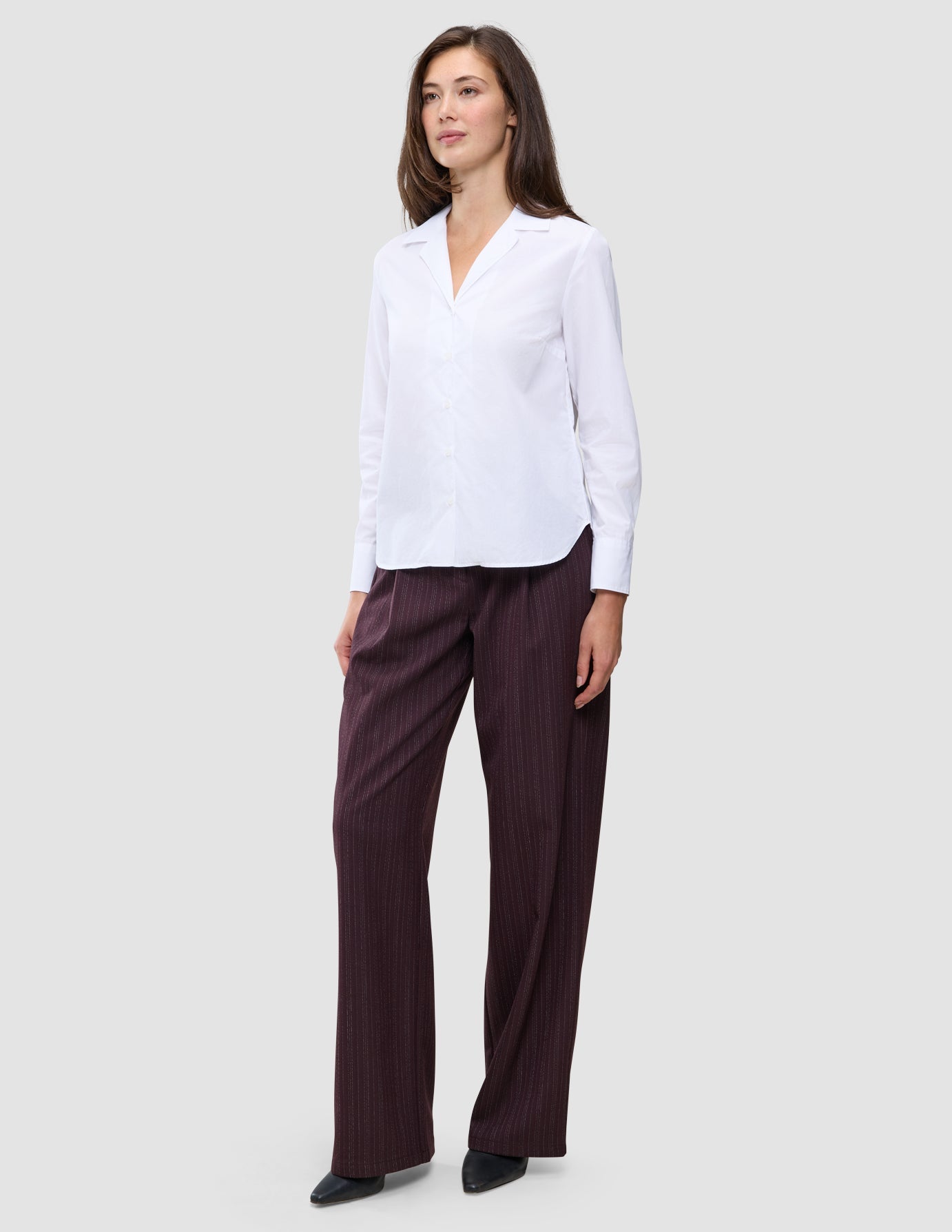A woman wears the white Madison Shirt featuring a notched collar, paired with loose-fitting dark pinstriped trousers.
