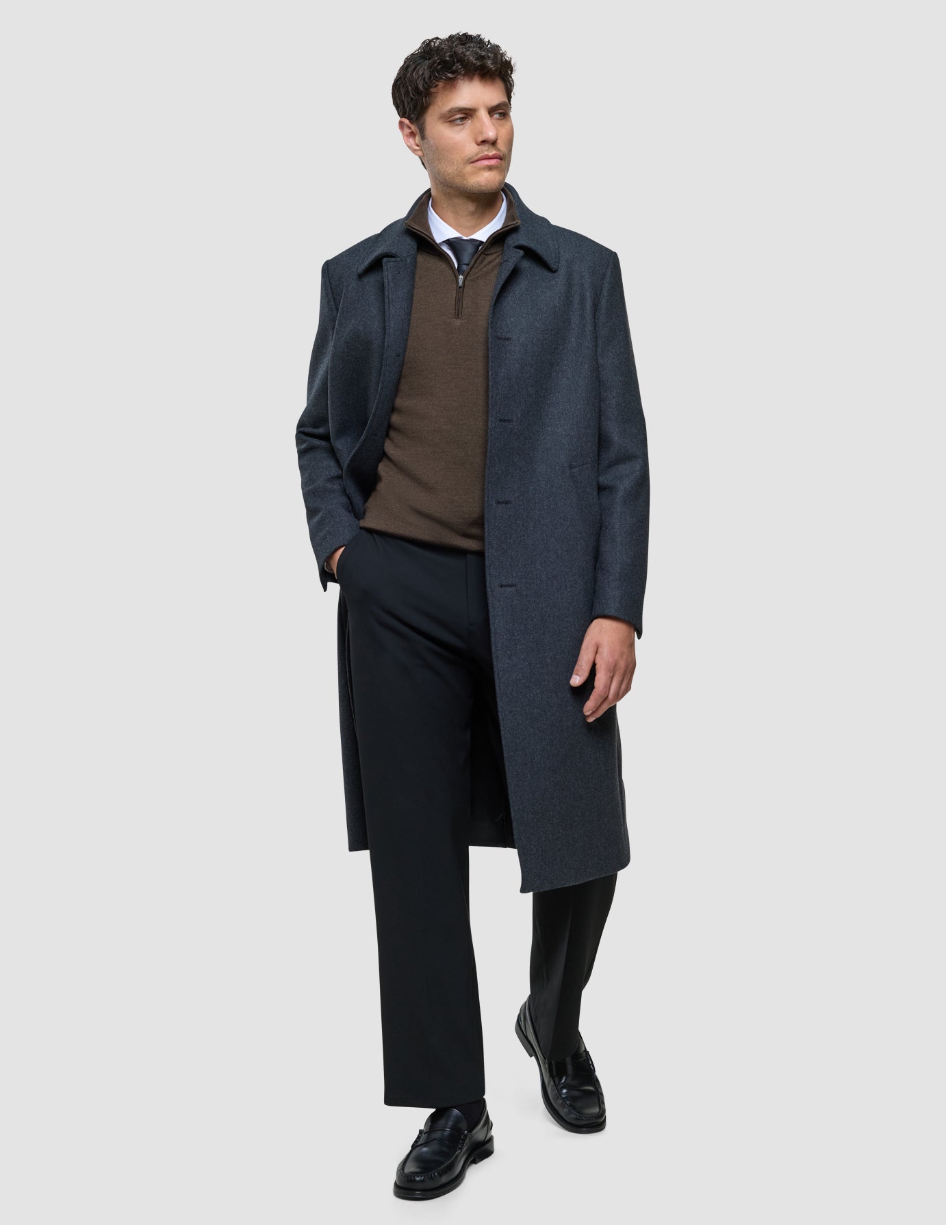 A man wears a Maxi Wool Coat Dark Grey over a brown quarter-zip sweater, a collared shirt, and black trousers.