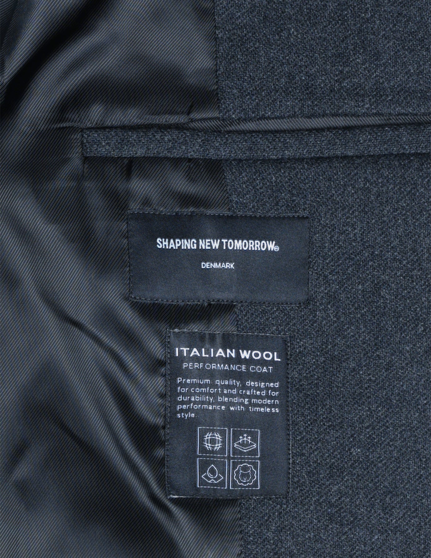 Interior labels of a dark grey Maxi Wool Coat from Shaping New Tomorrow, featuring text about the garment’s quality and style alongside four square icons.