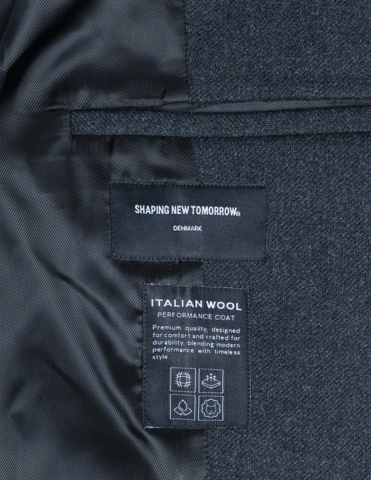 Interior labels of a dark grey Maxi Wool Coat from Shaping New Tomorrow, featuring text about the garment’s quality and style alongside four square icons.