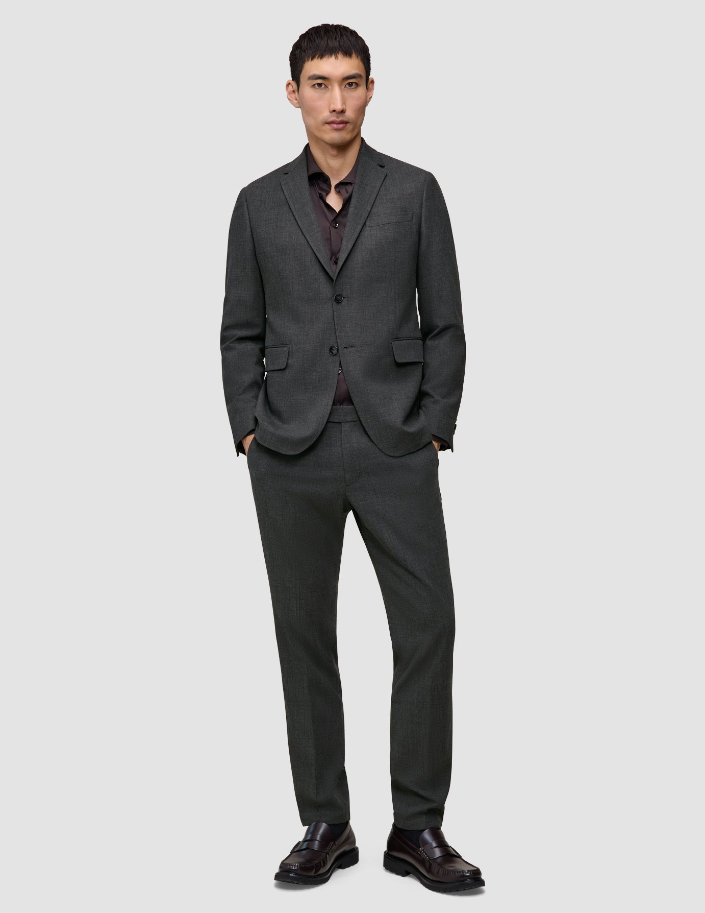 A man wearing a Motion Blazer Regular Pepper Grey and matching trousers over a dark button-down shirt, with his hands in his pockets.