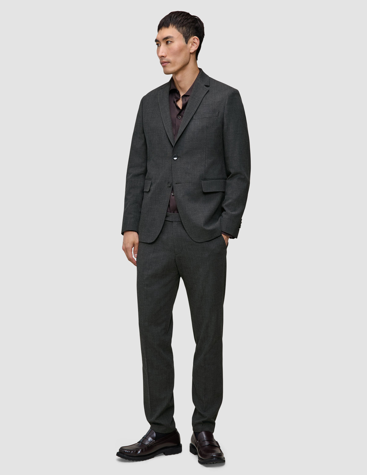 A man wearing a Motion Blazer Regular Pepper Grey tailored blazer and matching trousers over a dark brown button-down shirt and brown leather loafers.