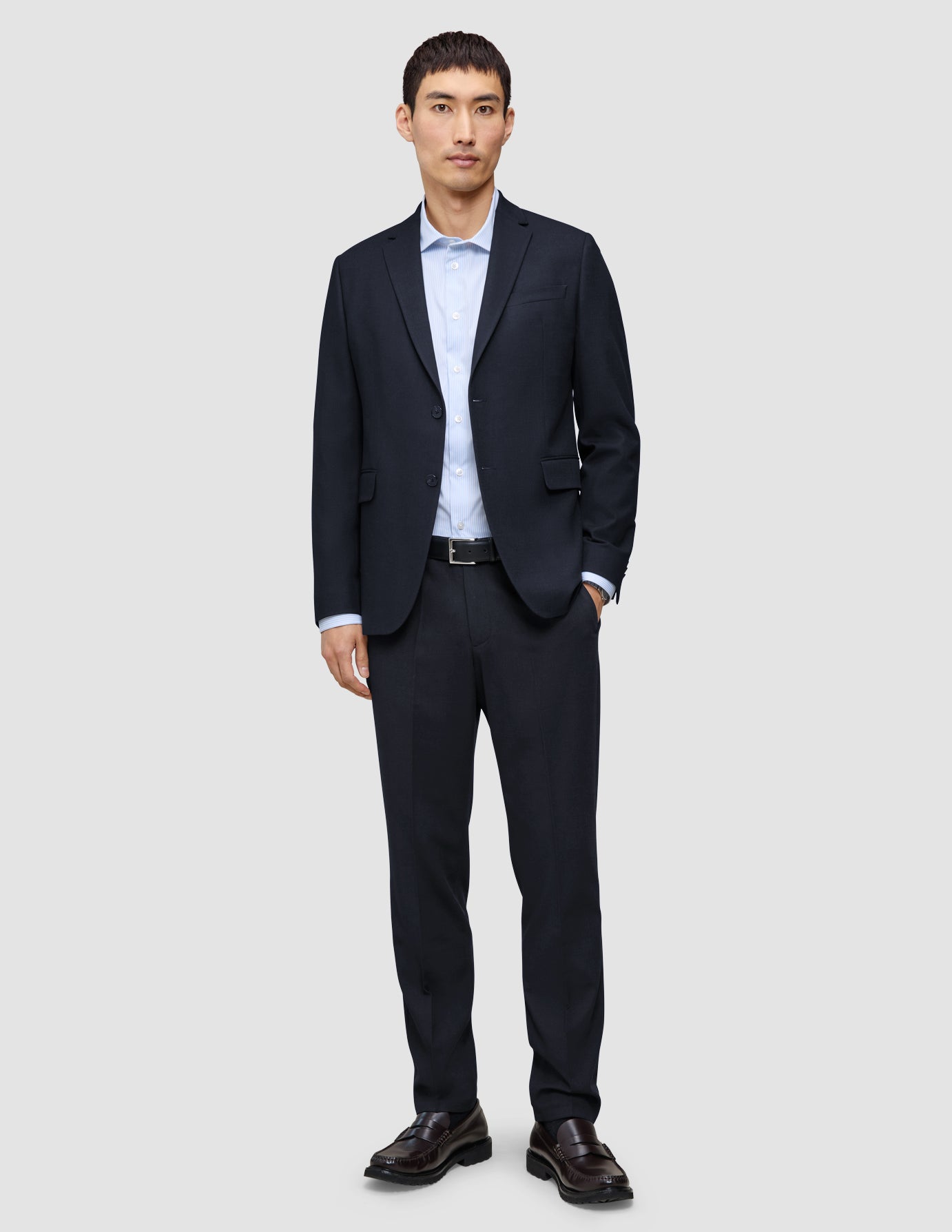 A man wearing a Motion Blazer Slim Midnight Blue with matching trousers, a light blue button-down shirt, a black belt, and brown loafers.