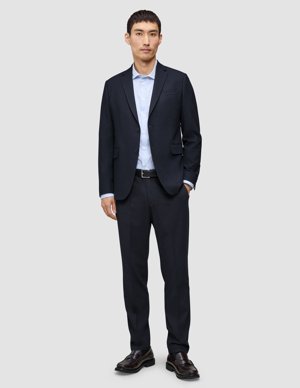 A man wearing a Motion Blazer Slim Midnight Blue with matching trousers, a light blue button-down shirt, a black belt, and brown loafers.