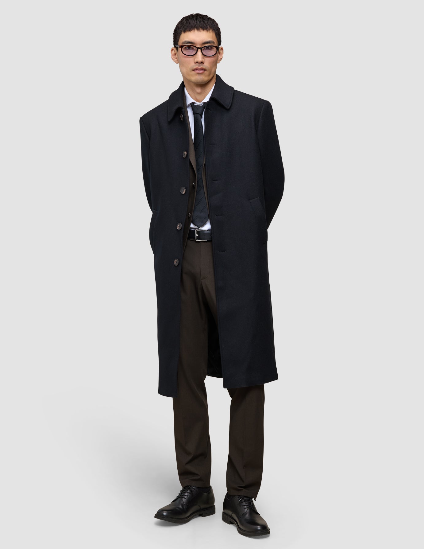 A man wears a black overcoat over a suit, white shirt, and tie, paired with Motion Suit Pants Regular Cognac and black dress shoes.