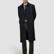 A man wears a black overcoat over a suit, white shirt, and tie, paired with Motion Suit Pants Regular Cognac and black dress shoes.