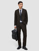A man wearing Motion Suit Pants Regular Cognac and a matching breathable jacket with a white dress shirt and dark tie, holding a black backpack.
