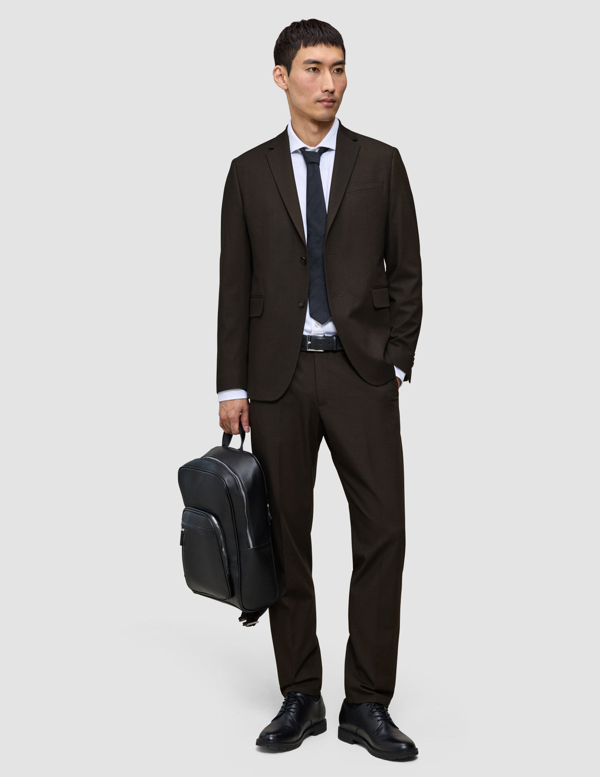 A man wearing Motion Suit Pants Regular Cognac and a matching breathable jacket with a white dress shirt and dark tie, holding a black backpack.