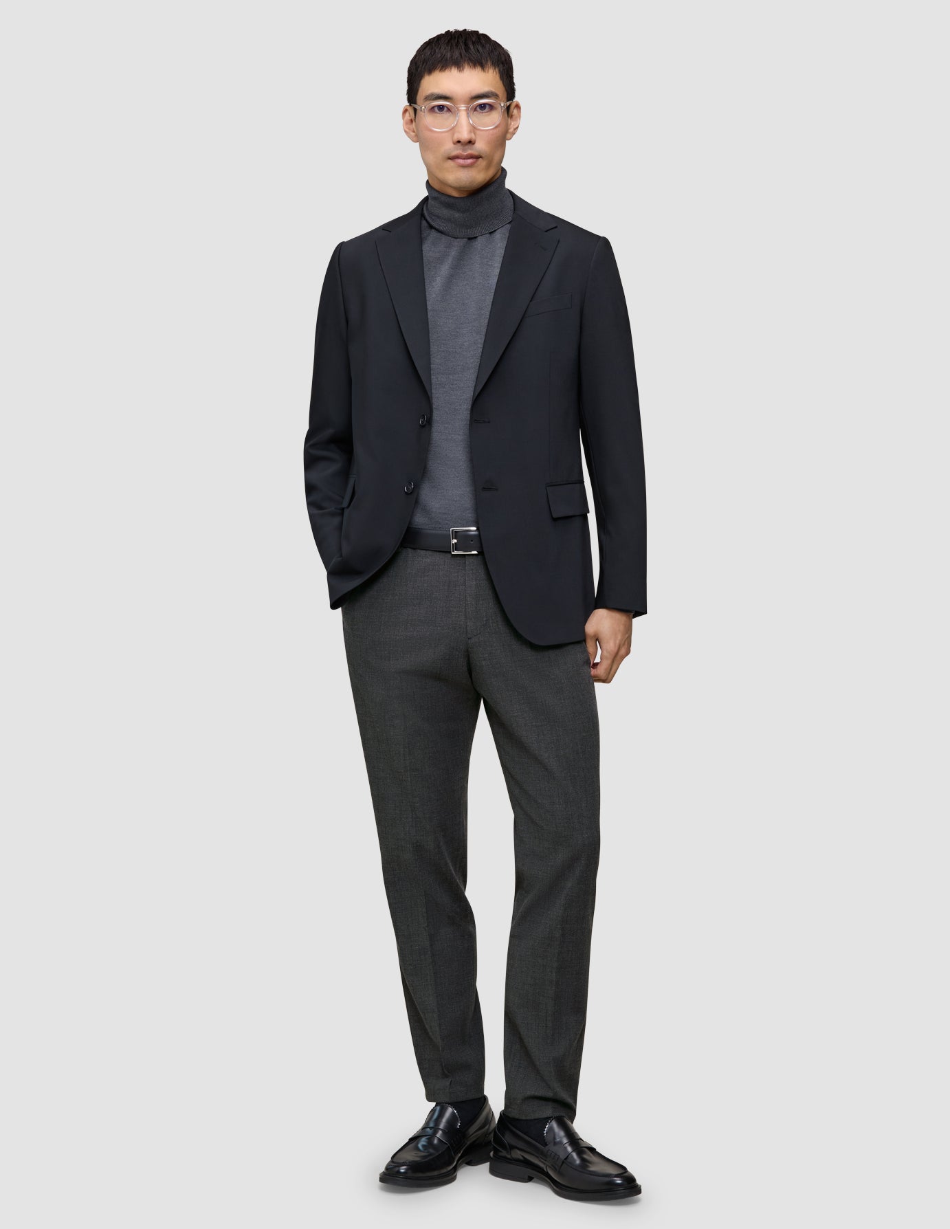 A man wears a black blazer, a grey turtleneck, and Motion Suit Pants Regular Pepper Grey, paired with a black leather belt and black loafers.