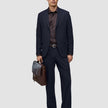 A man holding a brown leather briefcase, wearing a dark blue suit jacket, a dark brown button-down shirt, dark loafers, and Motion Suit Pants Relaxed Midnight Blue.