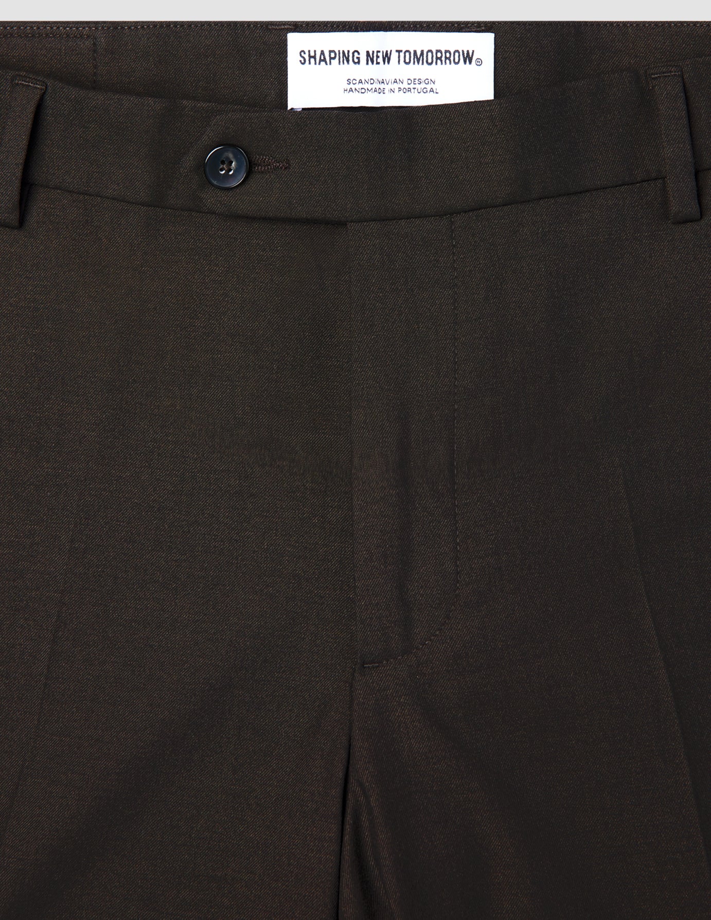 Close-up of Motion Suit Pants Slim Cognac in lightweight, breathable brown fabric, featuring a dark button and a brand label at the waistband.