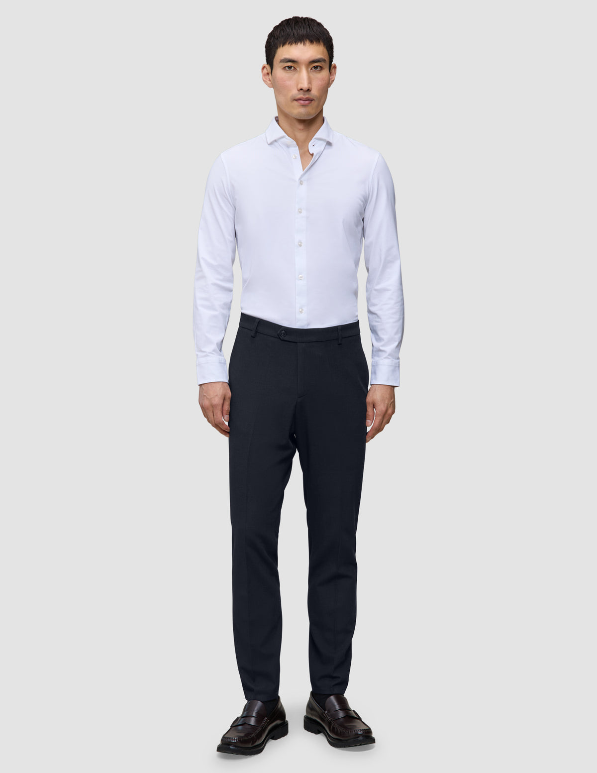A man wears a white button-down shirt, Motion Suit Pants Slim Midnight Blue, and brown loafers.