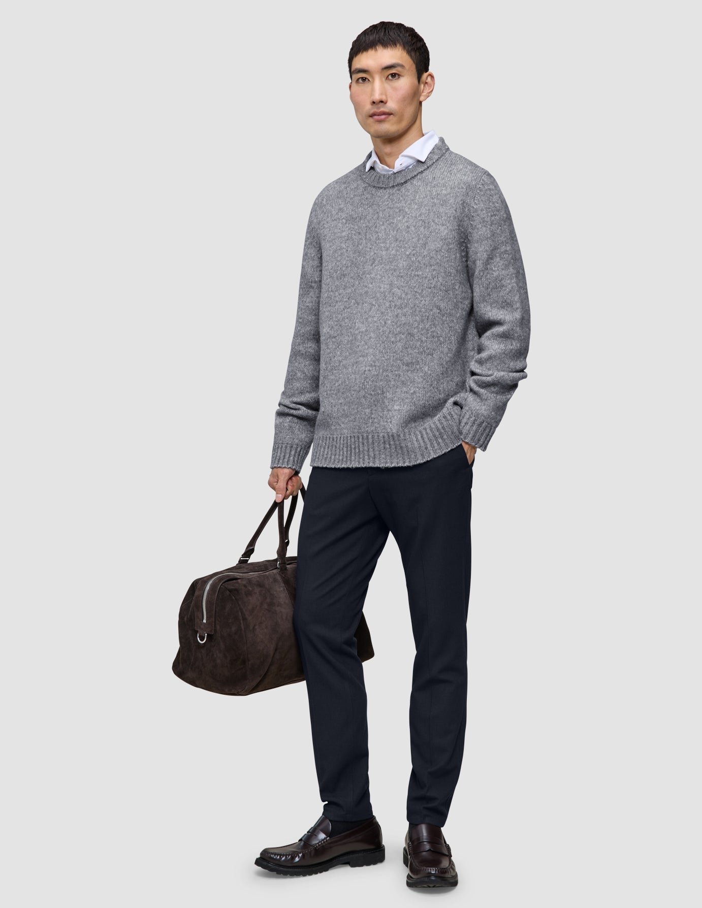 A man wearing a grey crewneck sweater over a white collared shirt and midnight blue Motion Suit Pants Slim, carrying a brown duffel bag and wearing dark loafers.
