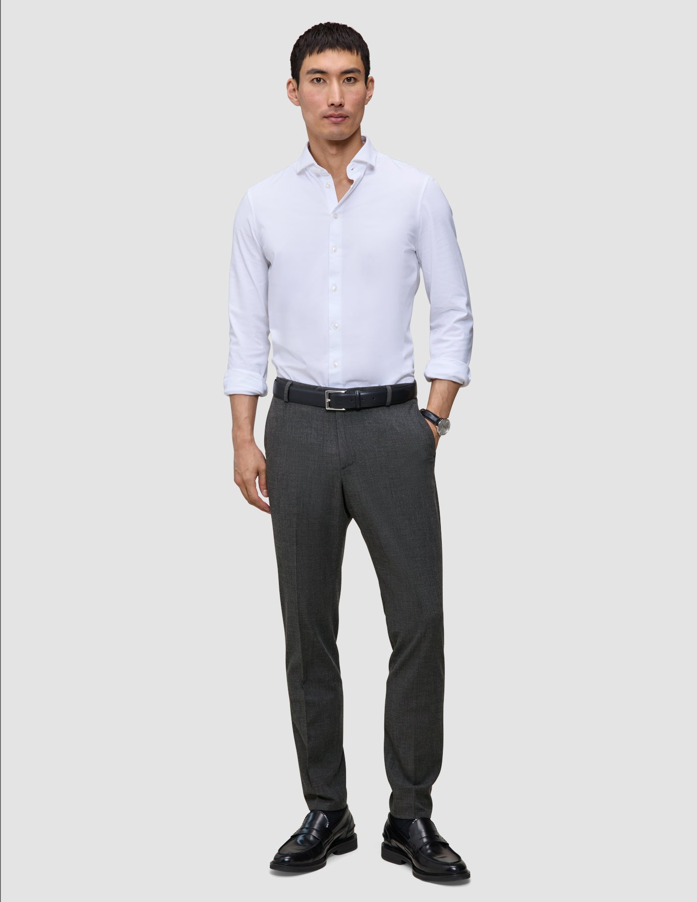 A man wearing a white button-down shirt, a black belt, and Motion Suit Pants Slim Pepper Grey made from breathable charcoal grey fabric.