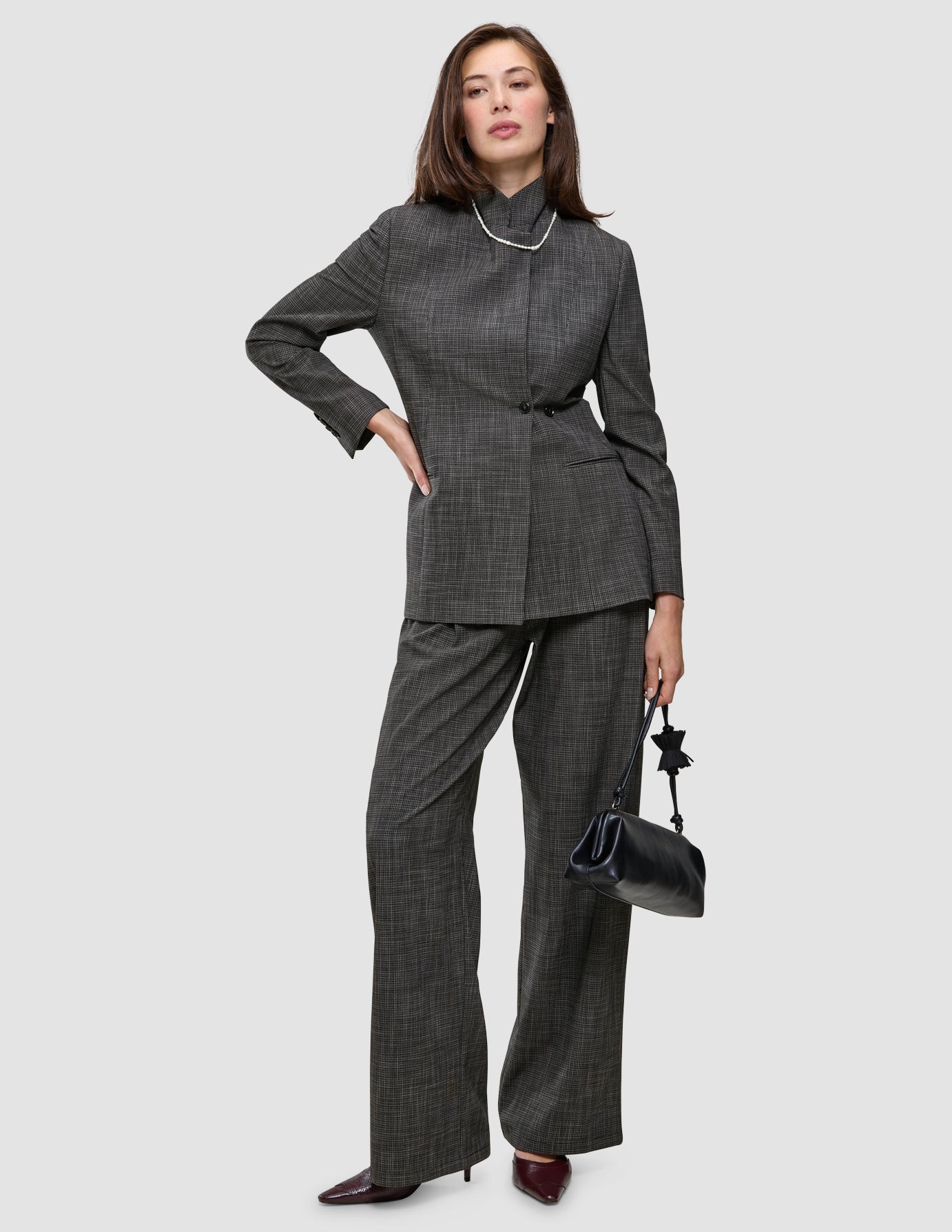 A person wearing The suit with a statement in a grey plaid Nara Blazer and wide-leg trousers, styled with a pearl necklace and a black handbag.