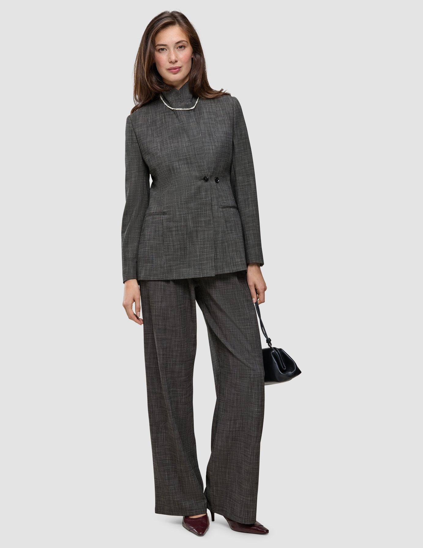 A woman wears The suit with a statement in grey plaid, featuring a blazer and matching wide-leg trousers, styled with a pearl necklace and a black handbag.