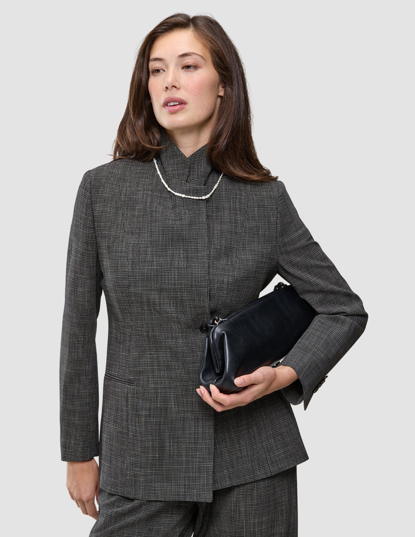 A woman wears a grey The suit with a statement blazer with matching wide-leg pants, a pearl necklace, and holds a black clutch.