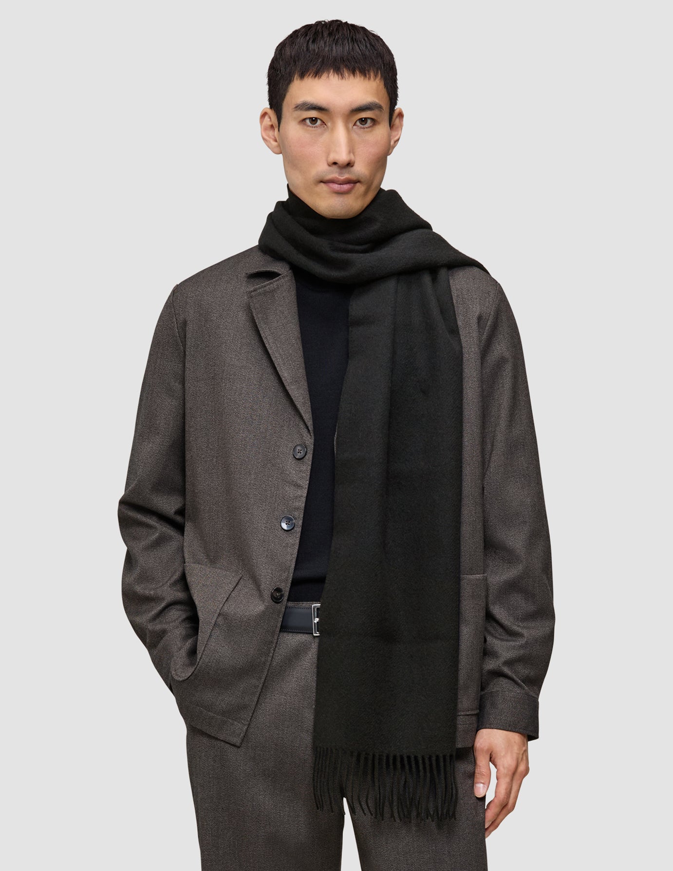 A person wearing a Scarf Black with tasseled ends over a black sweater and a brown jacket.