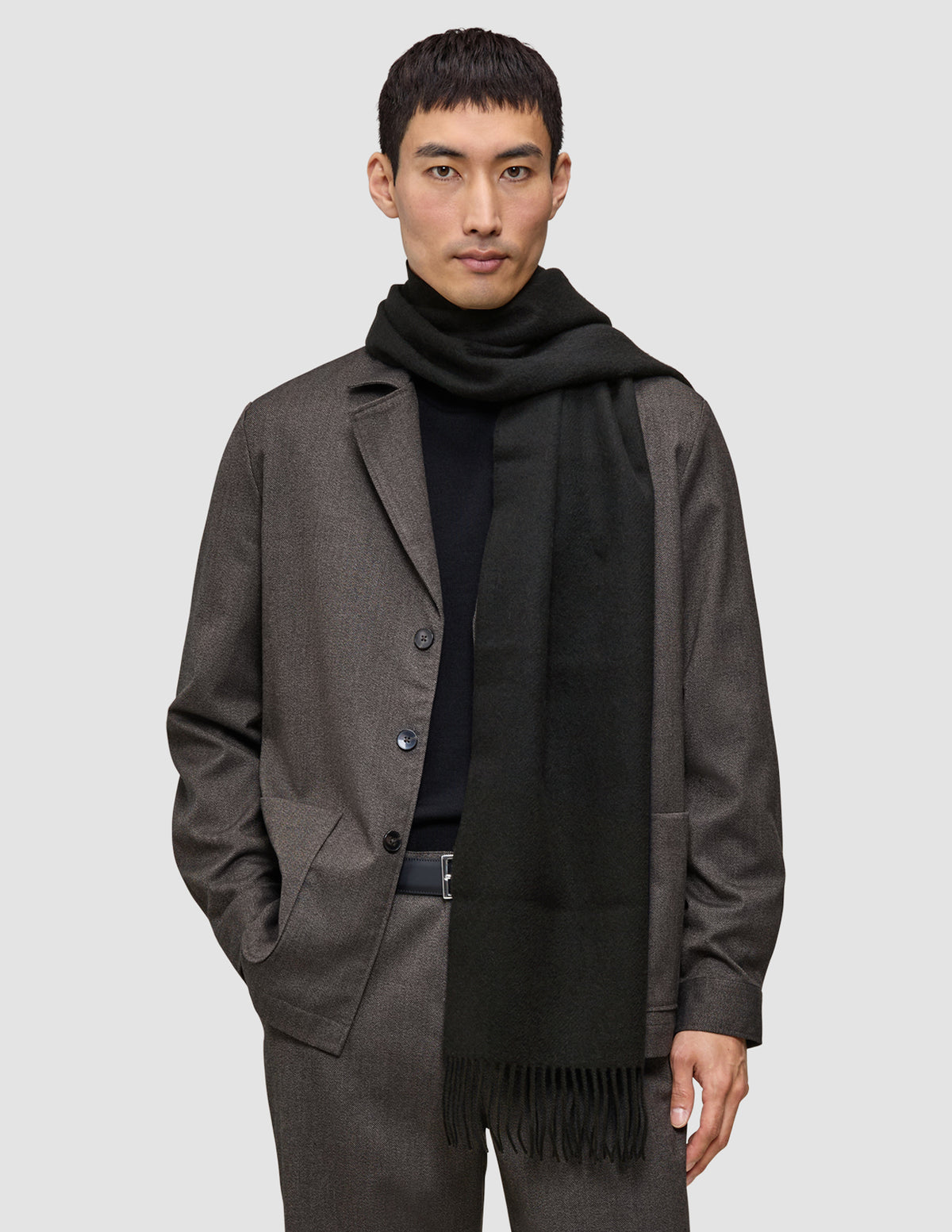 A person wearing a Scarf Black with tasseled ends over a black sweater and a brown jacket.