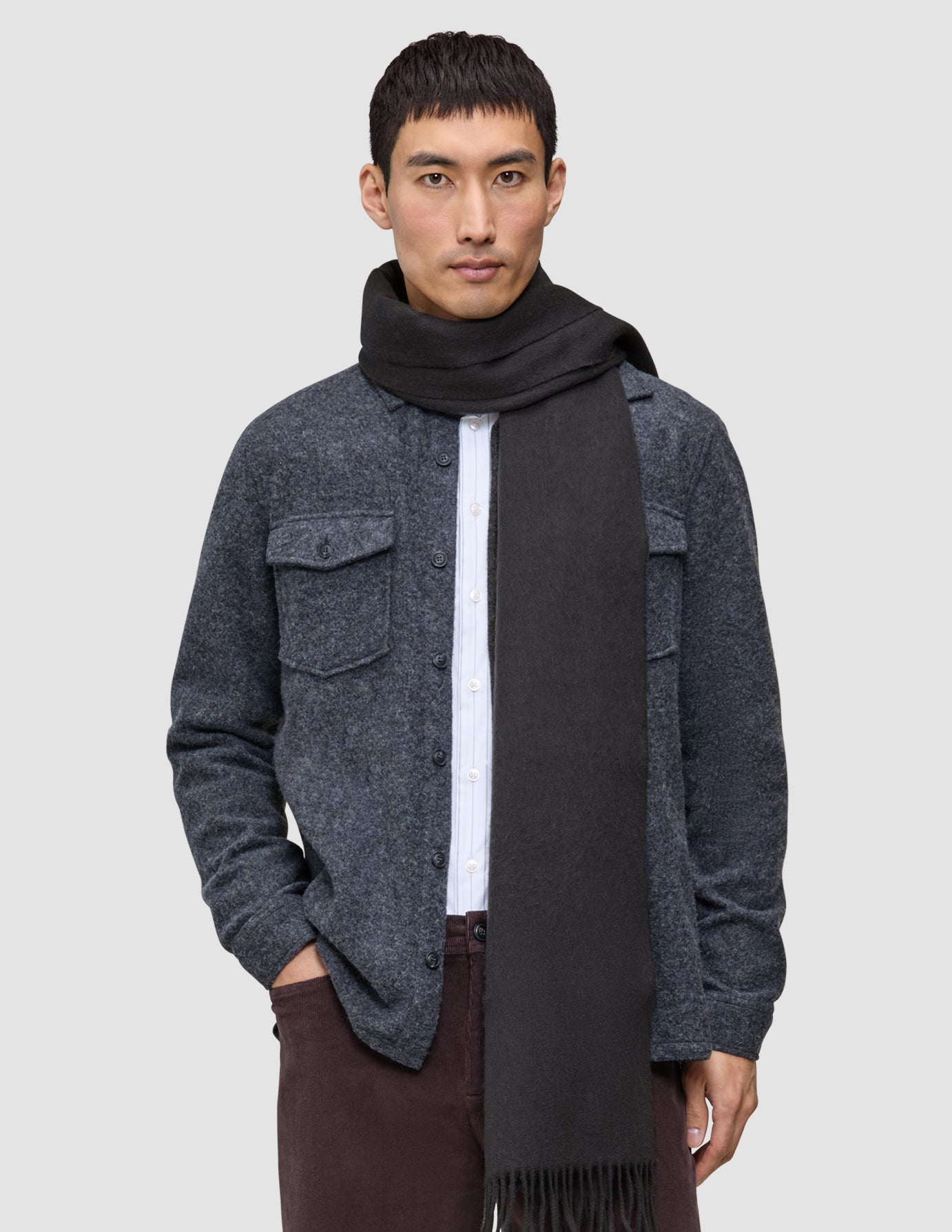 A man wears a Scarf Charcoal Grey with fringe ends over a white button-down shirt and a grey wool shirt jacket in a minimalist Scandinavian design.
