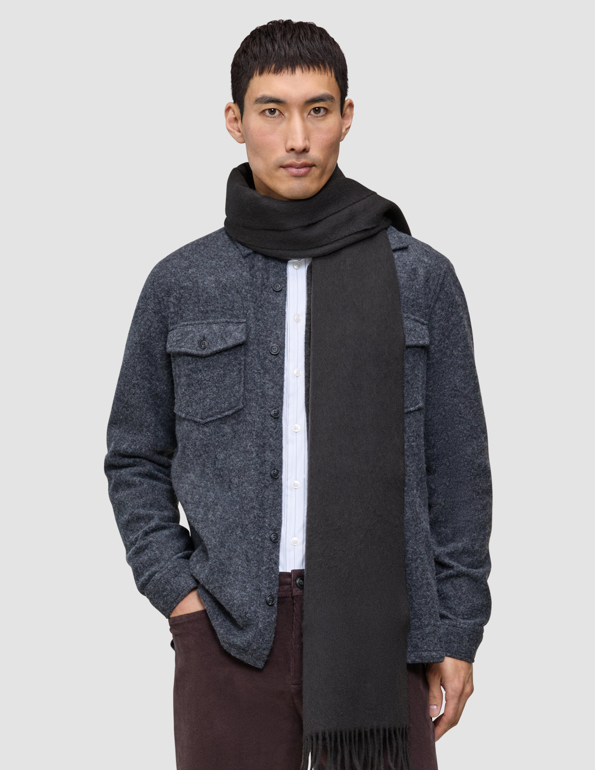 A man wears a Scarf Charcoal Grey with fringe ends over a white button-down shirt and a grey wool shirt jacket in a minimalist Scandinavian design.