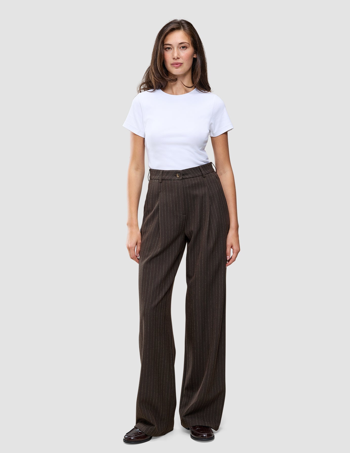 A person wearing a white t-shirt and Serene Pants Wide Walnut Herringbone, which are high-waisted, dark brown pinstriped trousers with a wide-leg silhouette.