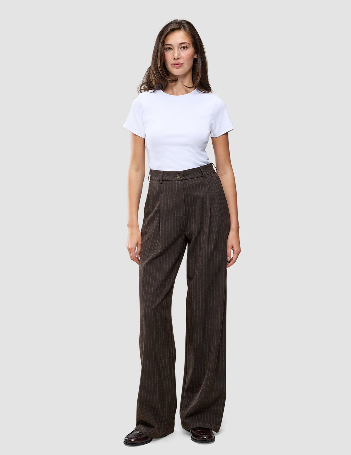 A person wearing a white t-shirt and Serene Pants Wide Walnut Herringbone, which are high-waisted, dark brown pinstriped trousers with a wide-leg silhouette.