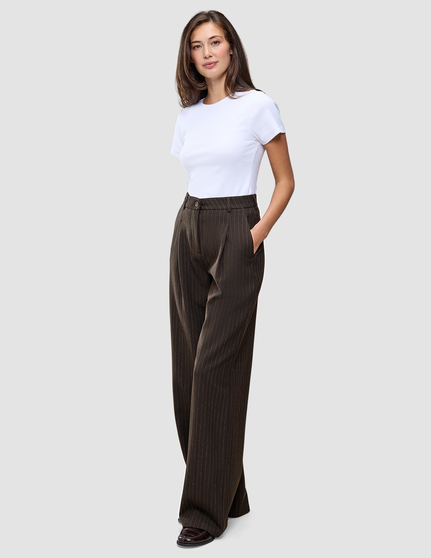 A woman stands wearing a white t-shirt and Serene Pants Wide Walnut Herringbone, which are dark, high-waisted wide-leg trousers with thin vertical pinstripes.