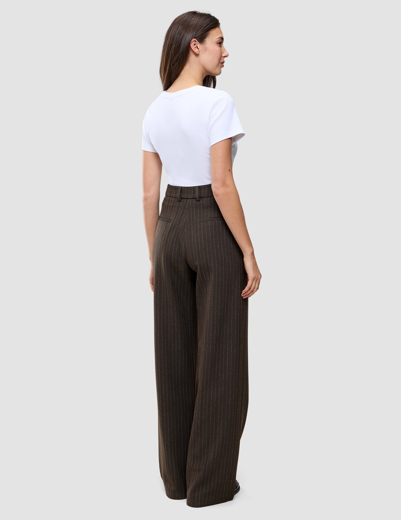 A woman wearing a white t-shirt and Serene Pants Wide Walnut Herringbone, high-waisted, dark brown, pinstriped, wide-leg trousers, viewed from the back.