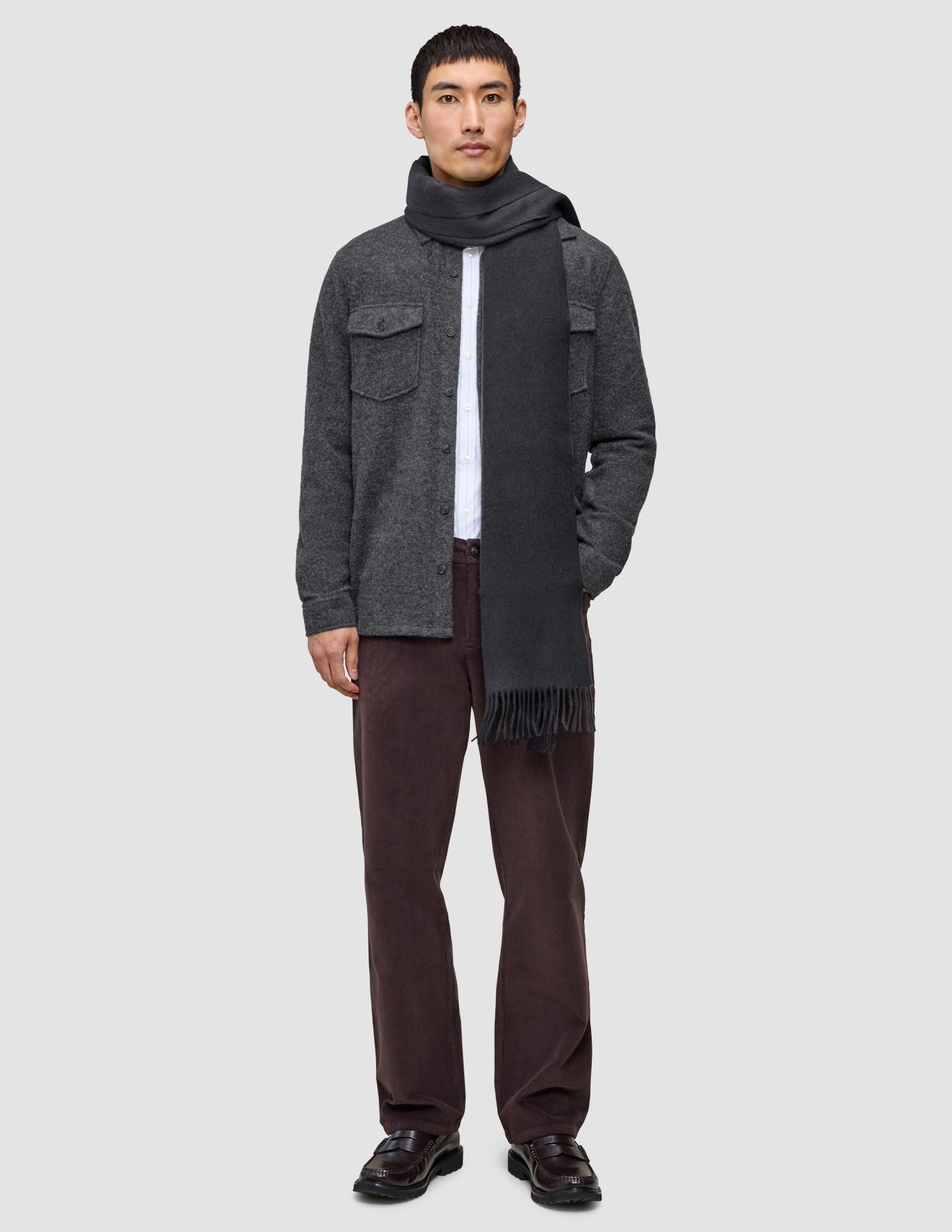 A man wearing a Casual and considered double layer wool overshirt over a white collared shirt, paired with a dark scarf, dark brown trousers, and loafers.