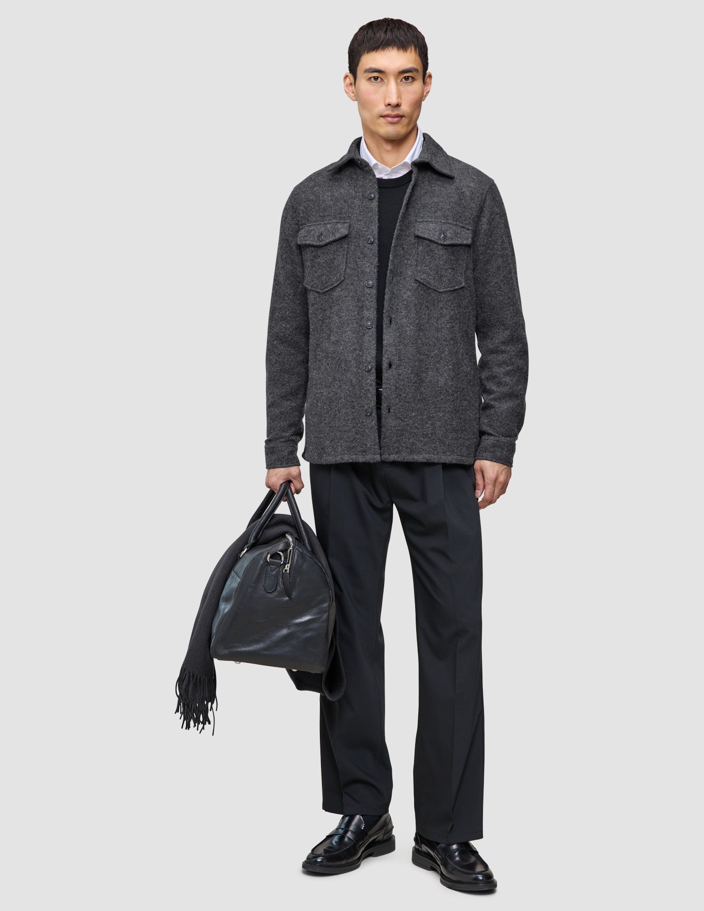 A man wearing Layers that work: a double layer wool overshirt over a white collared shirt and tech wool pants, holding a black leather duffel bag with a scarf draped over it.
