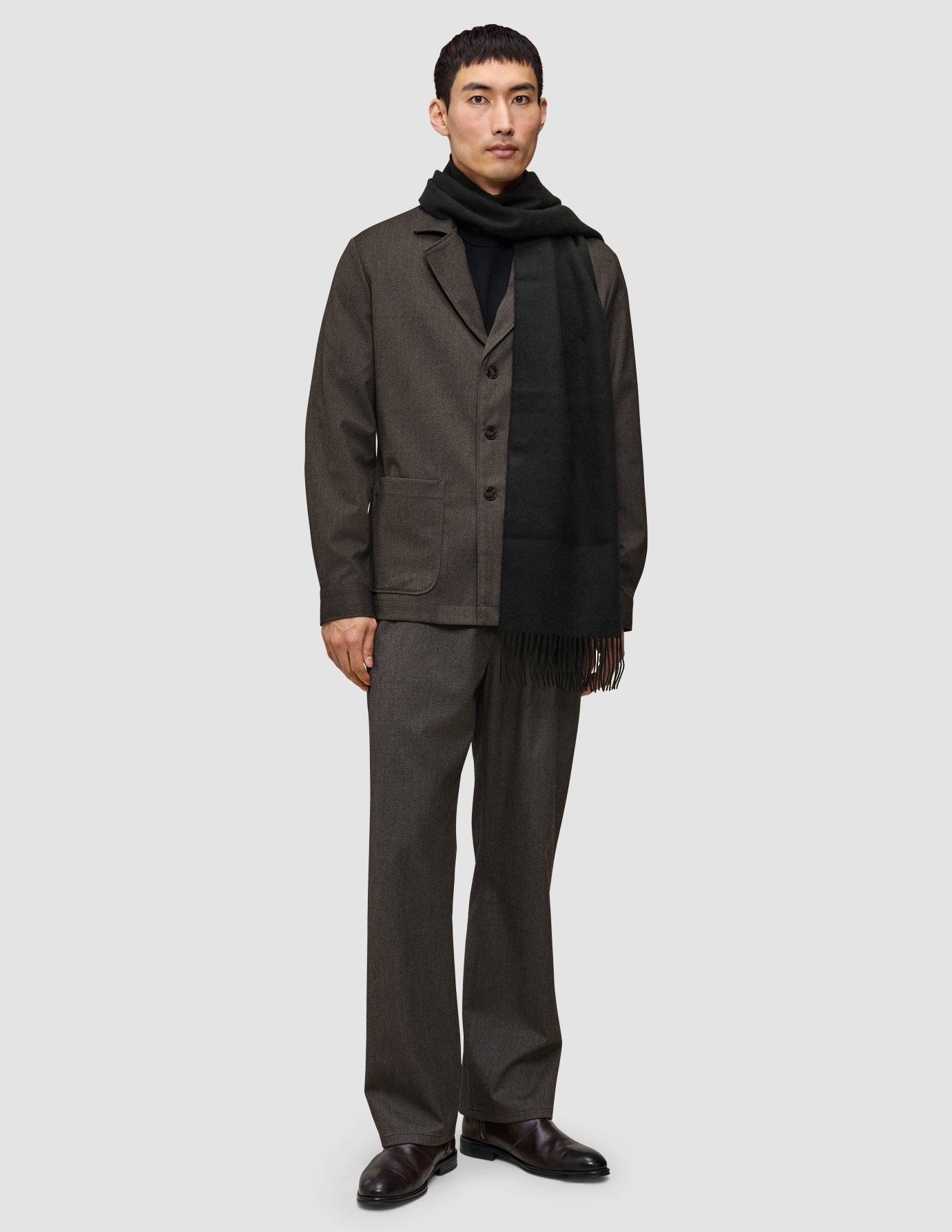 A man wearing a different kind of suit consisting of a dark brown Atelier Overshirt and matching Atelier Pants, styled with a black turtleneck, a long black scarf, and dark brown leather shoes.